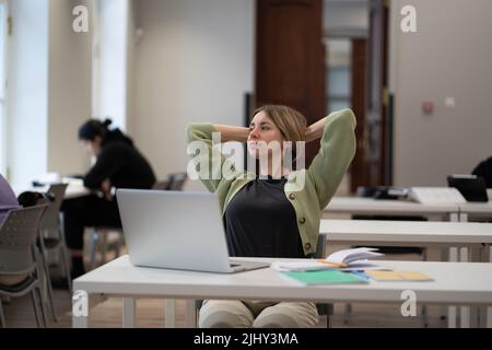 Verträumte Frau mittleren Alters, die sich mit den Händen über dem Kopf ausruht, während sie online in der Bibliothek studiert Stockfoto