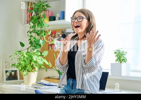 Positive lächelnde reife Frau, die zu Hause Sprachnachrichten am Telefon aufzeichnet Stockfoto