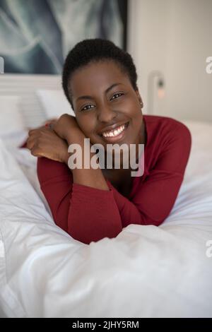 Nahaufnahme eines Porträts einer schönen schwarzafrikanischen Frau, die auf dem Bett liegt, mit gekreuzten Beinen und Armen, lächelnd und in die Kamera blickt Stockfoto