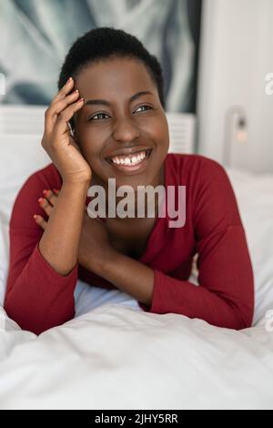 Nahaufnahme eines Porträts der schönen schwarzafrikanischen Frau, die auf dem Bett liegt, mit gekreuzten Beinen und Armen, lächelnd und wegblickend Stockfoto