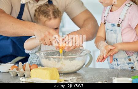 Vater knackt das Ei in eine Schüssel. Eltern backen mit seinen Kindern. Familienbacken in der Küche. Kaukasische Familie, die zusammen backt. Vater wird mit verbunden Stockfoto