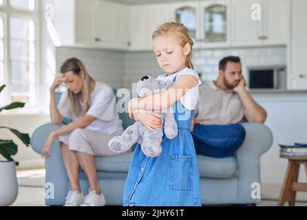 Ein verärgertes kleines Mädchen quetscht ihren Teddybär, während es traurig und deprimiert aussieht, während ihre Eltern im Hintergrund streiten. Denken Sie an ihre Eltern Stockfoto