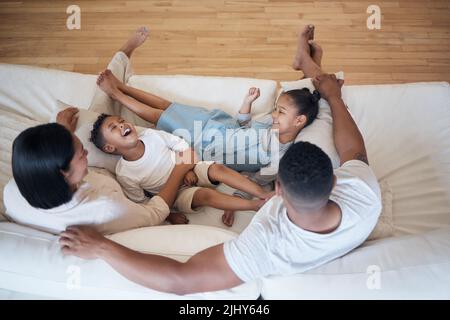 Fröhliche gemischte Rennfamilie aus vier Personen, die sich auf der Couch gemeinsam in der Lounge zu Hause entspannen. Fröhlicher Bruder und Schwester, die lachend Zeit mit den Eltern verbringen Stockfoto
