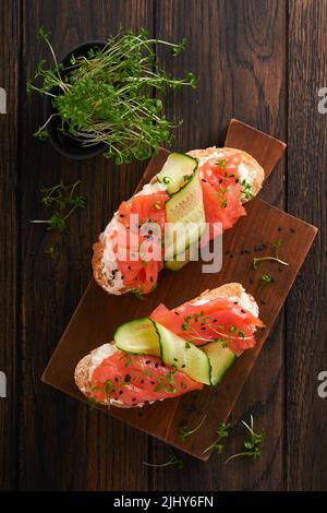 Sandwiches. Lachs-Toast mit Frischkäse, Gurke, schwarzem Sesam und Microgreens auf altem Holztisch-Hintergrund. Meeresfrüchte. Gesunde Ernährung. Fotografie Stockfoto