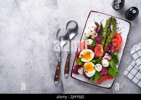 Spargel, Tomaten, Salat, Mozzarella, schwarzer Sesam, Flachs, Ölolivsalat und weich gekochtes Ei auf rechteckiger Keramikplatte auf grauem Betontisch Bac Stockfoto