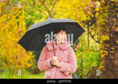 Glückliche junge asiatische Frau mit Regenschirm unter dem Regen im bunten Park Stockfoto