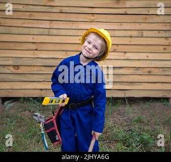 Niedlicher neugieriger Junge in einer blauen Overalls Uniform und einem Bauhelm mit Werkzeugen in den Händen. Spaß beim Lernen von Papa den Beruf eines BU Stockfoto