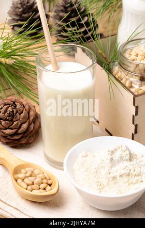 Ein Glas mit Zedernnussmilch, umgeben von Zapfen, Ästen, Nüssen, Zedernmehl in einer weißen Schüssel im Vordergrund. Gepulverte Alternativmilch. Gesund natürlich Stockfoto