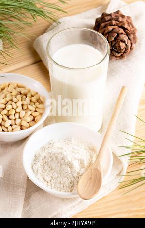 Glas Zedernnussmilch, Nüsse, Zedernmehl in weißer Schüssel mit Holzlöffel. Gepulverte Alternativmilch. Gesundes natürliches veganes Getränk. Mil. Auf Werksbasis Stockfoto