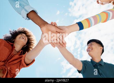 Eine niedrige Nahaufnahme einer Gruppe von verschiedenen jungen Freunden, die sich an einem sonnigen Tag mit einem blauen Himmel im Hintergrund die Hände in einem Huddle zusammenschließen Stockfoto
