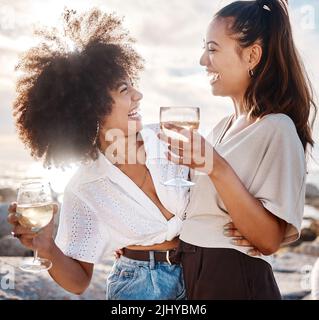 Zwei junge, gemischte Rassen-Freundinnen, die am Strand rumhängen und Wein trinken. Hispanische Frauen, die draußen ein Glas Wein trinken Stockfoto