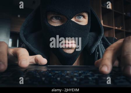 Ein Mann in einer Maske druckt auf der Tastatur bei Ein Tisch im Dunkeln Stockfoto