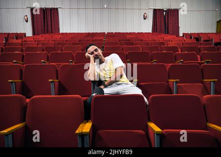 Junger Mann mit Brille schläft allein im Kinosaal, während alle weg sind. Ein einsamer Mann in einem leeren Theater schlief bei der Vorstellung ein. Ein Unint Stockfoto