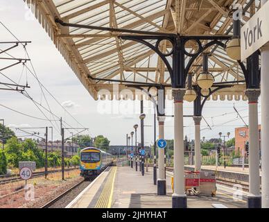 Ein Zug fährt von einem Bahnhof ab. Es passiert das Ende einer Plattform mit einem historischen Vordach. Ein Himmel mit Wolken ist oben. Stockfoto