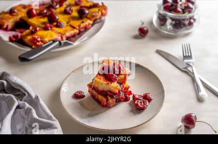 Süßer Kirschkuchen oder roter Brownie-Kuchen. Ein Stück Kuchen auf dem Teller, ganzer Kuchen und rohe Kirschen im Rücken Stockfoto