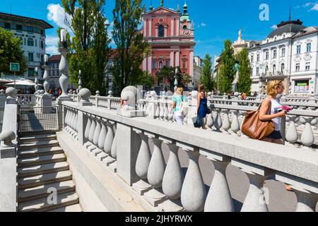 Fußgänger auf der Tromostovje (Dreifachbrücke) über den Fluss Llubljanica. Franziskanerkirche der Verkündigung im Hintergrund. Ljubljana, Slowenien. Stockfoto