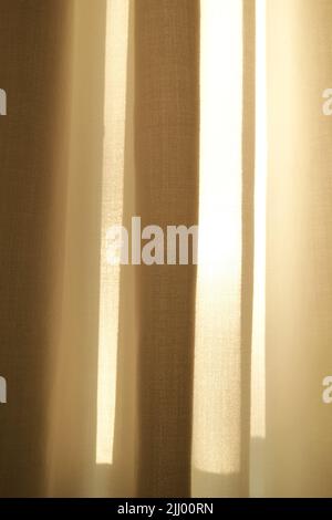 Geschlossene Leinenvorhänge, die an einem sonnigen Tag in einem modernen Haus am Fenster hängen. Schatten und Sonnenlicht, die durch cremefarbene Baumwolle oder beigefarbene Stoffe leuchten Stockfoto