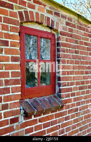 Altes, dreckiges Fenster in einem roten Backsteinhaus. Antiker Flügel mit rotem Holzrahmen in einem historischen Gebäude mit klumpiger Farbstruktur. Äußere Details eines Stockfoto