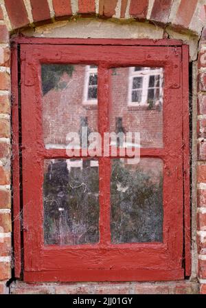 Nahaufnahme eines alten schmutzigen Fensters in einem roten Backsteinhaus. Zoomen Sie auf den Flügel mit rotem Holzrahmen in einem historischen Gebäude mit holpriger Farbstruktur. Makro Stockfoto