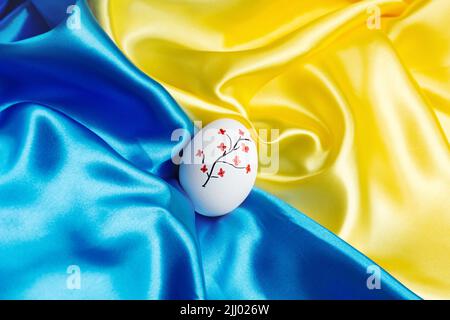 Weißes Osterei auf der blauen und gelben Staatseidenflagge der Unabhängigkeit der Ukraine. Ein Baum mit roten Blumen. Alles gute zum Tag der Unabhängigkeit und der Verfassung. Frohe Ostern und Feiertage. Stockfoto