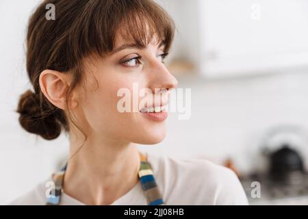 Weiße junge Frau mit Schürze lächelnd und schaut zu Hause zur Seite Stockfoto