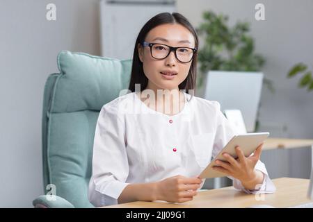 Online-Aufnahme ins Krankenhaus. Eine junge, schöne asiatische Ärztin zeichnet die Krankengeschichte eines Patienten online auf einer Tablette auf. Tippen Sie auf ein Tablet. Sitzen am Tisch im Büro in der Winding. Stockfoto