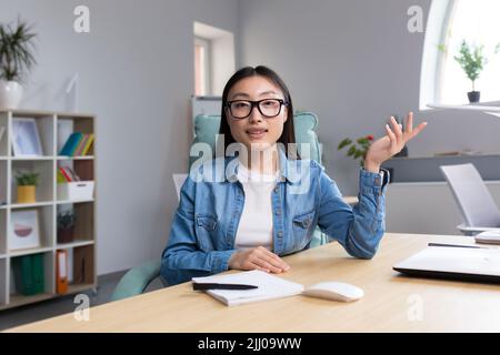 Fernstudium. Eine junge Asiatin ist Lehrerin, führt Online-Kurse, Unterricht durch. Er schaut in die Kamera, erklärt, erzählt. Sitzen in Brille und Jeans an einem Tisch an einem Computer in einem Klassenzimmer Stockfoto