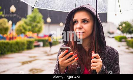 Junge, schöne Frau, die auf der Straße der Stadt mit einem transparenten Regenschirm im Regen auf dem Handy steht. Ziemlich weiblich mit Smartphone-Schutz mit Stockfoto