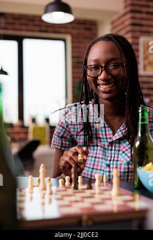 Glückliche Frau, die beim Schachspielen Pfand in der Hand hatte und herzlichst vor der Kamera lächelte. Ein clever aussehender Brettspielspieler hält ein Schachbrett und denkt über den nächsten Schritt nach, während er seine Freizeit genießen kann. Stockfoto