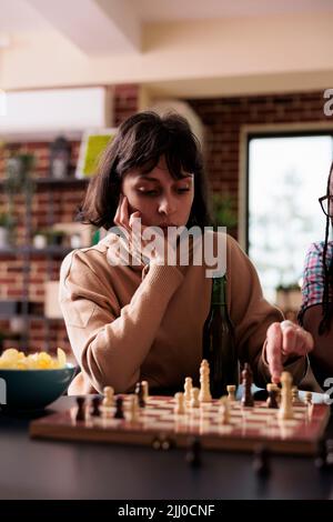 Nachdenkliche Frau, die am Tisch im Wohnzimmer sitzt, während sie über den nächsten Schachzug nachdenkt. Intelligenter Brettspielspieler, der ein Stück auf dem Schachbrett bewegt, während er sich zu Hause mit Freunden entspannt, die Spaß haben. Stockfoto