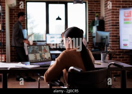 Frau mit chronischer Beeinträchtigung nimmt an Videocall-Treffen in einem behindertengerechten Büro Teil, chattet mit Menschen auf Online-Videokonferenzen. Rollstuhlfahrer sprechen über Fernkonferenz. Stockfoto