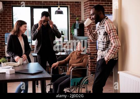 Ein vielseitiges Team von Kollegen, die im Geschäftsbüro lachen und eine gelähmte Frau im Rollstuhl sitzen. Asiatischer Mitarbeiter mit chronischer Behinderung und Beeinträchtigung, der Spaß mit Kollegen hat. Stockfoto