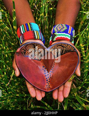 Die Hände in bunten Armbändern halten einen herzförmigen Teller Stockfoto