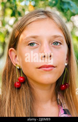 Defocus schöne blonde Teenager-Mädchen mit süßen Kirsche auf dem Ohr. Schöne lächelnde Teenager-Mädchen Nahaufnahme auf dem Grün des Sommerparks. Ohrringe für Kinder. Sommer por Stockfoto