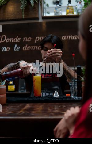 Latino Barkeeper bereitet vor einem Mädchen in einem roten Kleid ein gelbes Getränk zu. Kellner an der Bar, der ein Getränk zubereitete. Mädchen wartet auf einen Drink, während sie es zubereiten. Kellner im Nachtclub. Stockfoto