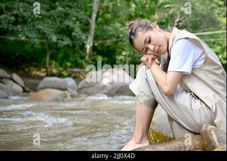 Ruhige und entspannte, tausendjährige asiatische Frau, die auf dem Felsen in der Nähe des Flusses im wunderschönen Wald sitzt. Trekking und Camping Outdoor-Aktivitätskonzept. Stockfoto