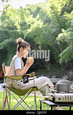 Ruhige und entspannte junge asiatische Frau, die auf dem Campingplatz campt, auf einem Campingstuhl sitzt, einen Kaffeebecher hält und am Morgen Kaffee schlürft. Stockfoto