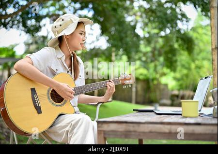 Entspannte und glückliche junge asiatische Frau singt ein Lied, während sie während ihrer Urlaubspause im schönen Campinggarten Gitarre spielt. Aktivitäten im Freien CO Stockfoto