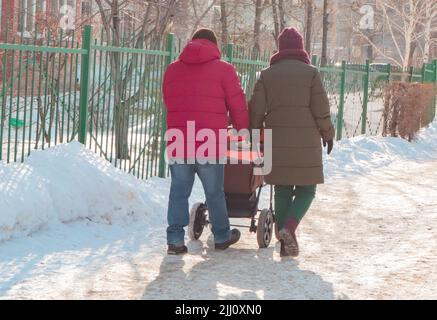 Ein Paar mit einem Kinderwagen spaziert während eines Spaziergangs durch den Winterpark. Schneewetter, das Konzept einer kompletten traditionellen Familie Stockfoto