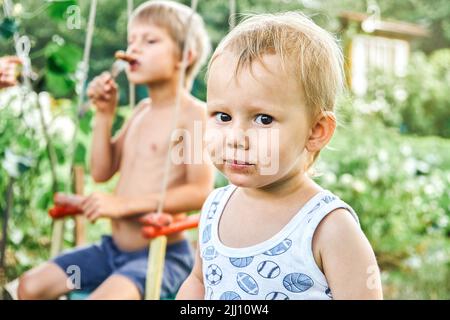 Brüder genießen Grillwurst bei einem Picknick in der Natur mit üppigem Grün in der Landschaft. Porträt von niedlichen blonden Kleinkind Junge auf verschwommenem Hintergrund Stockfoto