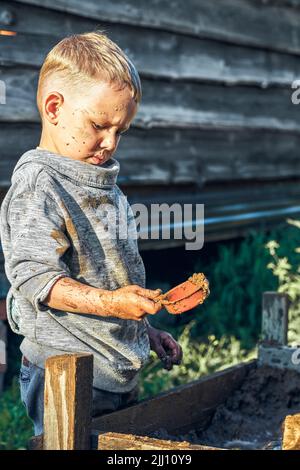 Nette ernsthafte Vorschulkinder mit schmutzigem Gesicht und Kleidung, die mit Spielzeugautos im Sandkasten in der Nähe von hölzernen ländlichen Haus am Sommertag auf dem Land spielt Stockfoto