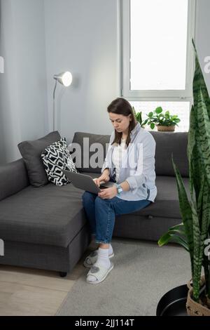 Frau mittleren Alters, die von zu Hause aus arbeitet. Mädchen mit Laptop zu Hause, auf dem Sofa sitzend. Remote-Work-Konzept Stockfoto