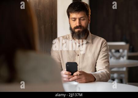 Kollegen von Mann und Frau trinken Kaffee im Café, Mann mit Smartphone sitzt in der Kaffeepause am Tisch, keine Freizeit, Hintergrund Stockfoto