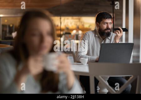 Bärtiger Geschäftsmann, der im Café arbeitet, verwendet einen Sprachassistenten für Laptop und Mobiltelefon, ein verschwommenes Mädchen im Vordergrund, einen selektiven Fokus Stockfoto