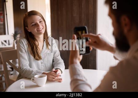 Verliebtes Paar sitzt im Café, männlicher Fotograf fotografiert Frau auf dem mobilen Smartphone, Indoor-Handy-Fotografie, sieht liebevoll Freund oder Stockfoto