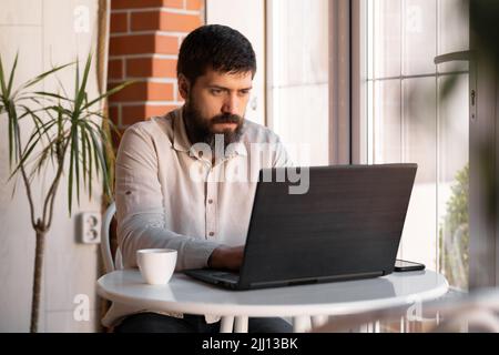 Gutaussehender bärtiger Mann, der im Restaurant am Laptop arbeitet, ein Mann, der im Online-Freiberufler arbeitet, trinkt Kaffee in einem Café Stockfoto