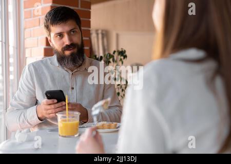 Glückliches junges Paar, das im Café frühstückte und sich mit dem Handy amückte. Café oder Restaurant Interieur. Kopierraum Stockfoto