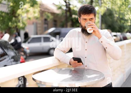 Mann, der Kaffee zum Mitnehmen trinkt, steht auf der Straße mit dem Mobiltelefon, bärtiger Mann, der im Straßencafé im sozialen Netzwerk chattet Stockfoto