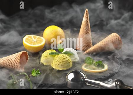 Leckeres cremiges und zitroniges Eis mit Minze, das auf einem schwarzen Hintergrund auf einem Steinschiefer in Rauch serviert wird. Stockfoto