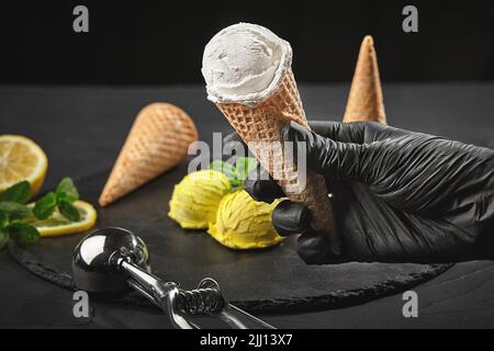 Hand in einem dunklen Handschuh hält einen Waffelkegel mit einem cremigen Eis auf dem Hintergrund eines mit Minze dekorierten Zitronensorbets. Stockfoto